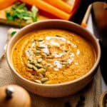 A bowl of creamy Air Fryer Vegan Carrot Soup topped with pumpkin seeds and a swirl of cream, with carrot sticks and fresh herbs in the background.