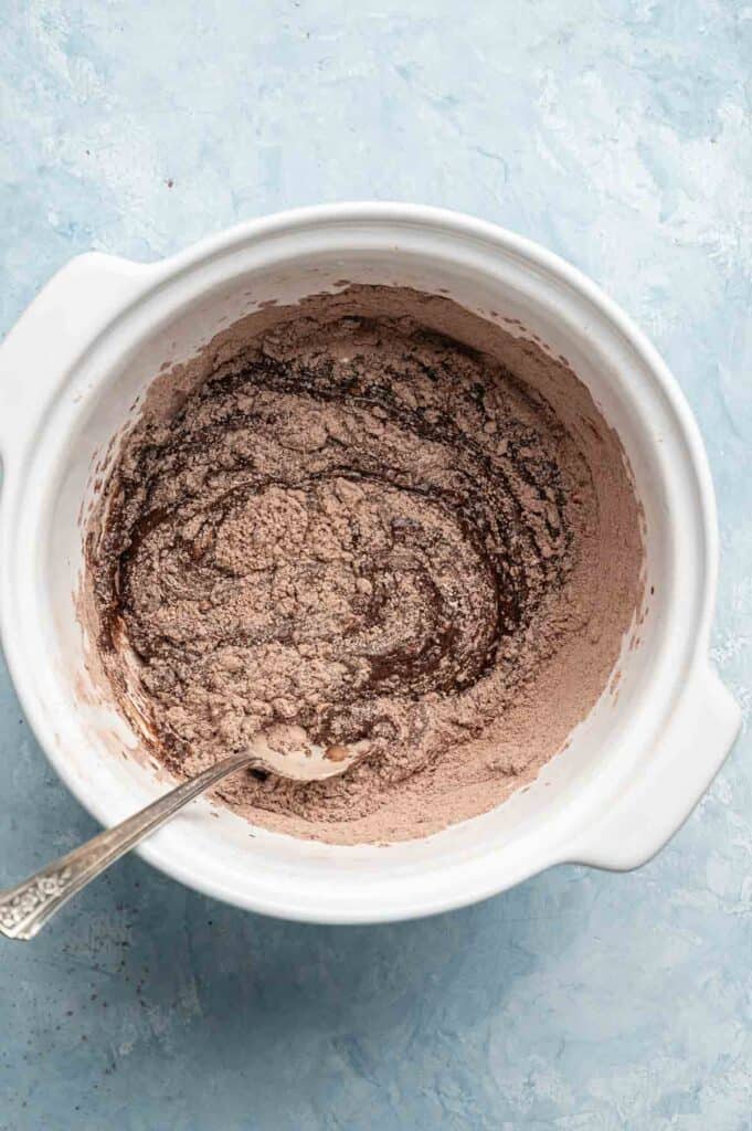 A white mixing bowl contains partially mixed chocolate batter with dry ingredients visible, ready to be transformed into delicious Air Fryer Vegan Crinkle Cookies, with a metal spoon resting inside.