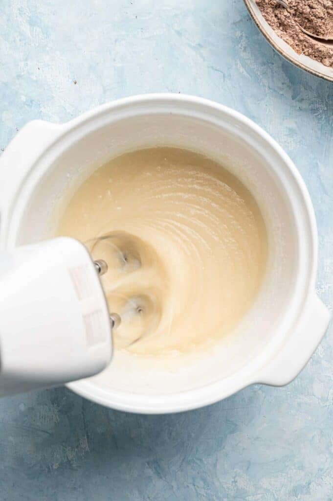 An electric hand mixer blends a creamy batter in a white mixing bowl on a light blue surface, preparing the base for Air Fryer Vegan Crinkle Cookies, with a bowl of dry ingredients partially visible nearby.