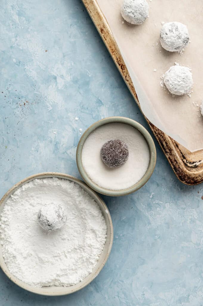 Two bowls with sugar and powdered sugar; one chocolate dough ball in each bowl, partially coated. Four coated dough balls rest on a parchment-lined baking sheet—perfectly prepped to become delicious Air Fryer Vegan Crinkle Cookies. Light blue background.