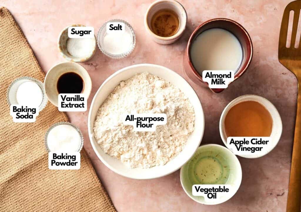 Top-down view of labeled baking ingredients in bowls on a pink surface, perfect for making Air Fryer Vegan Pancakes, including flour, sugar, salt, almond milk, apple cider vinegar, oil, vanilla, baking soda, and baking powder.