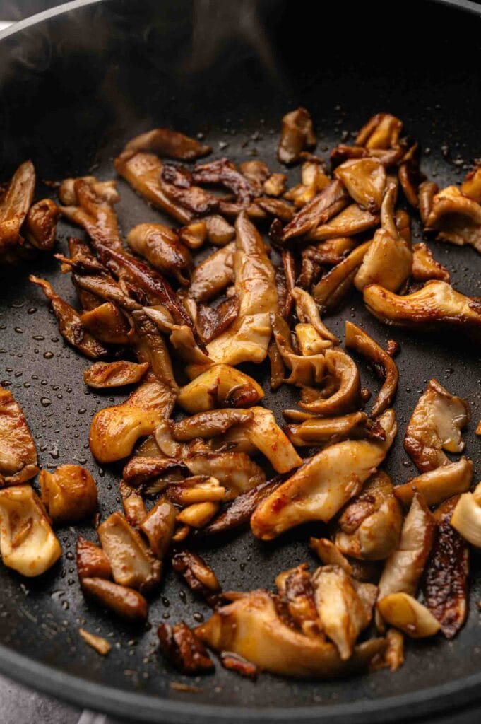 Sliced mushrooms are saut&eacute;ed in a black frying pan, showing a golden-brown color and a light sheen from cooking oil.