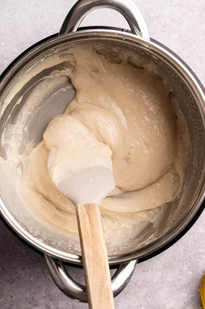 A spatula stirs a thick, creamy white mixture in a stainless steel pot on a light gray surface, preparing the perfect filling for Vegan Quesadillas with Mushroom Bacon.