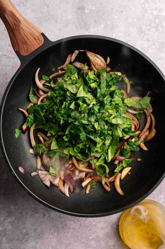Chopped spinach and sliced red onions are sautéing in a black frying pan, with a small bottle of olive oil nearby on a gray countertop—perfectly prepared as flavorful fillings for Vegan Quesadillas with Mushroom Bacon.