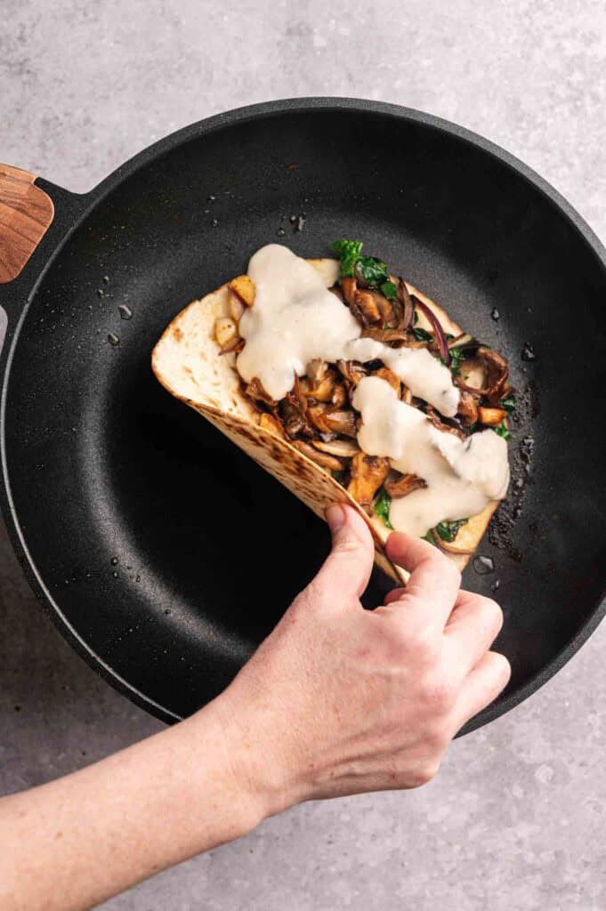 A hand folds a tortilla filled with mushrooms, greens, and melted cheese in a black skillet on a gray surface. 