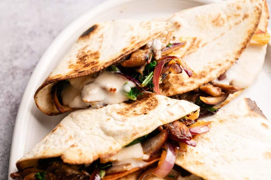 Three folded Vegan Quesadillas with Mushroom Bacon, saut&eacute;ed red onions, herbs, and melted cheese are served on a white plate.