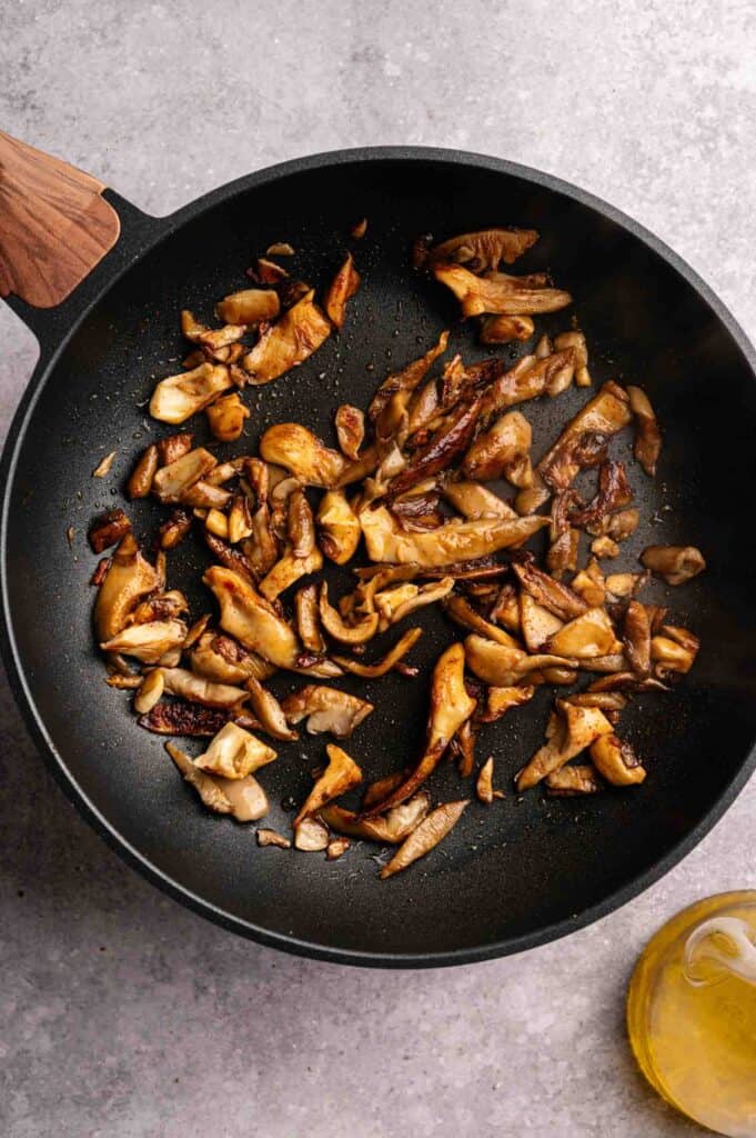Sliced mushrooms, perfect for Vegan Quesadillas with Mushroom Bacon, sauté in a black skillet with a wooden handle, placed on a gray surface. A small glass container of oil sits nearby.