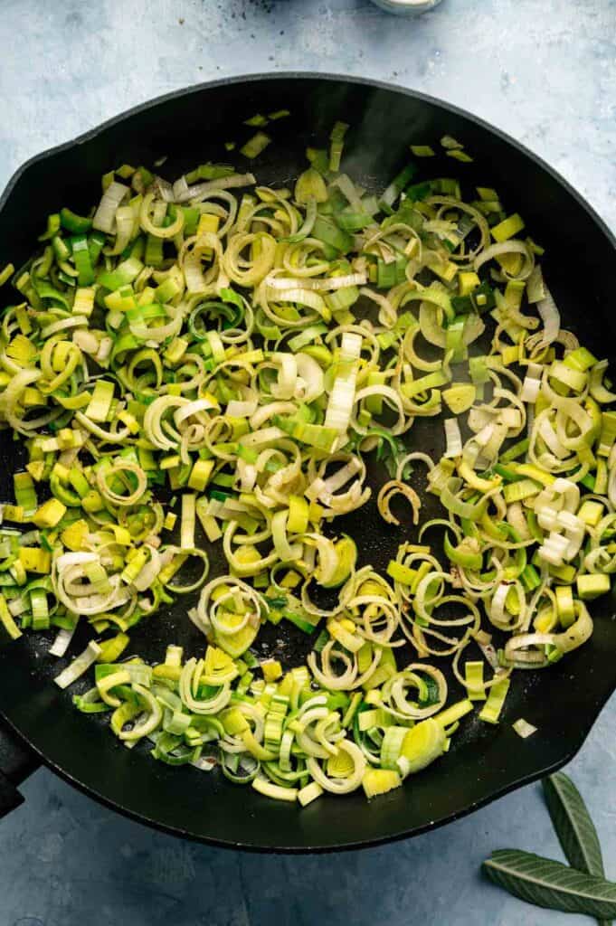 Sliced leeks cooking in a black skillet on a light blue surface, with steam rising from the vegetables, create a vibrant base for an Air Fryer Vegan Squash Casserole.