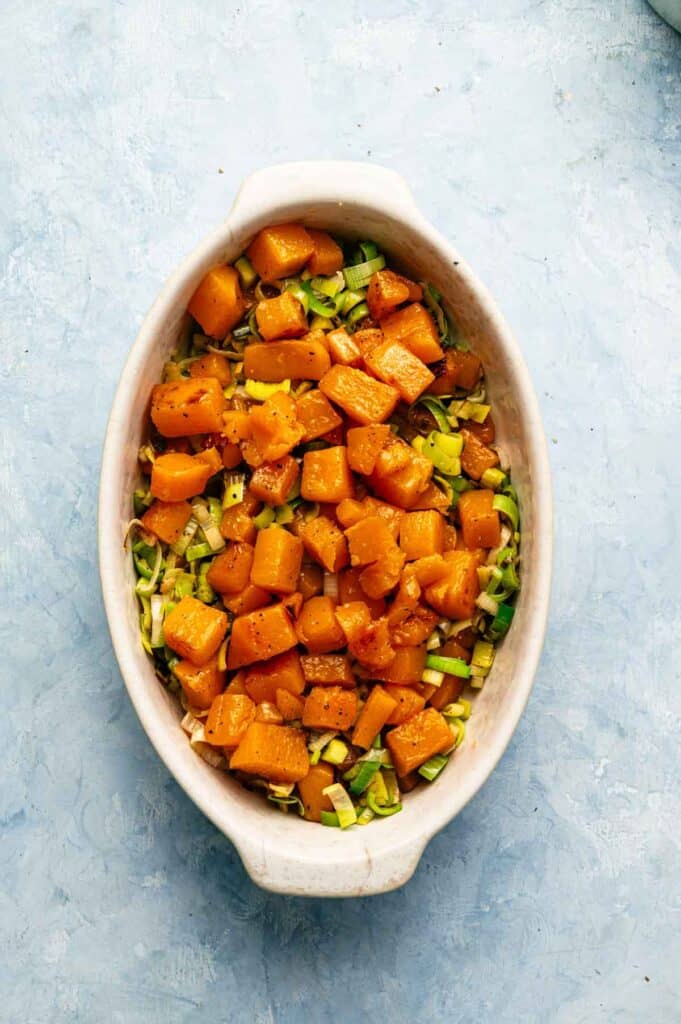 Oval dish filled with roasted butternut squash cubes layered over chopped green vegetables on a light blue textured surface, this Air Fryer Vegan Squash Casserole is both vibrant and wholesome.