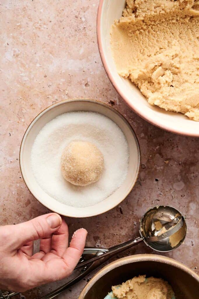 A hand rolls a ball of Air Fryer Vegan Sugar Cookies dough in a bowl of sugar, with extra dough and a metal scoop nearby on the countertop.