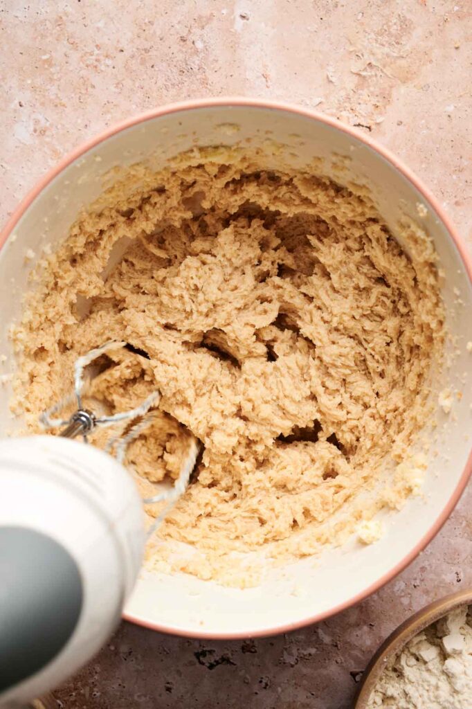 A mixing bowl filled with Air Fryer Vegan Sugar Cookies dough being mixed with a hand mixer on a light-colored countertop.