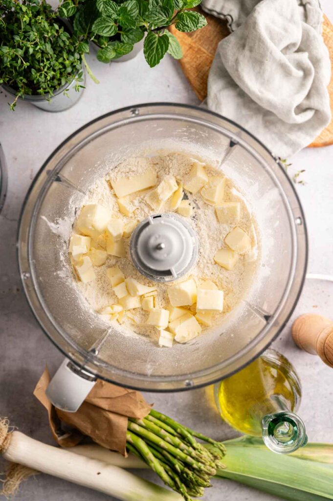 Food processor bowl filled with flour and cubed butter, surrounded by fresh herbs, asparagus, a leek, olive oil, and a kitchen towel&mdash;perfect for prepping an Air Fryer Vegan Asparagus Quiche on your countertop.