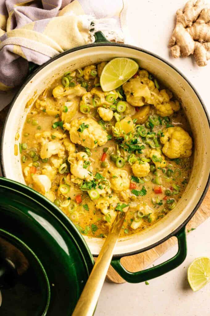 A pot of coconut curry with cauliflower, green onions, and herbs, garnished with lime, sits on a countertop with a cloth and fresh ginger nearby.