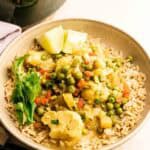 A bowl of a vibrant coconut curry featuring peas, carrots, cauliflower, and potatoes, garnished with cilantro and lime wedges.