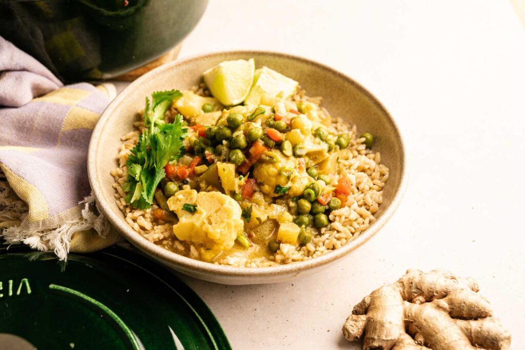 A bowl of coconut curry featuring peas, carrots, and potatoes, garnished with cilantro and lime wedges. Fresh ginger and a cloth are nearby.