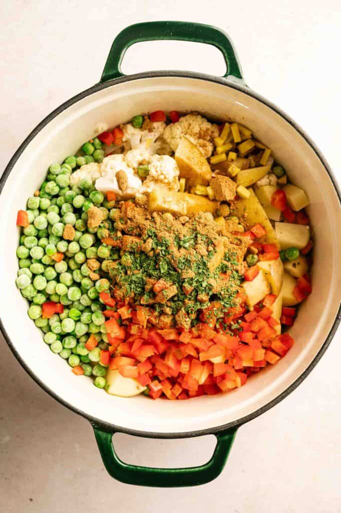 A pot filled with chopped vegetables including peas, cauliflower, potatoes, carrots, and bell peppers, topped with spices and herbs, ready to be cooked into a flavorful coconut curry.