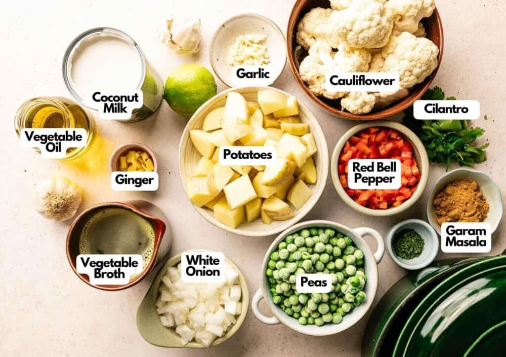 Various ingredients for a coconut curry are arranged in bowls, including potatoes, cauliflower, peas, red bell pepper, white onion, garlic, ginger, cilantro, garam masala, coconut milk, and vegetable broth.