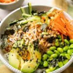 A vibrant Sushi Bowl featuring rice topped with sliced avocado, shredded carrots, cucumber, edamame, seaweed pieces, black sesame seeds, and a creamy sauce.