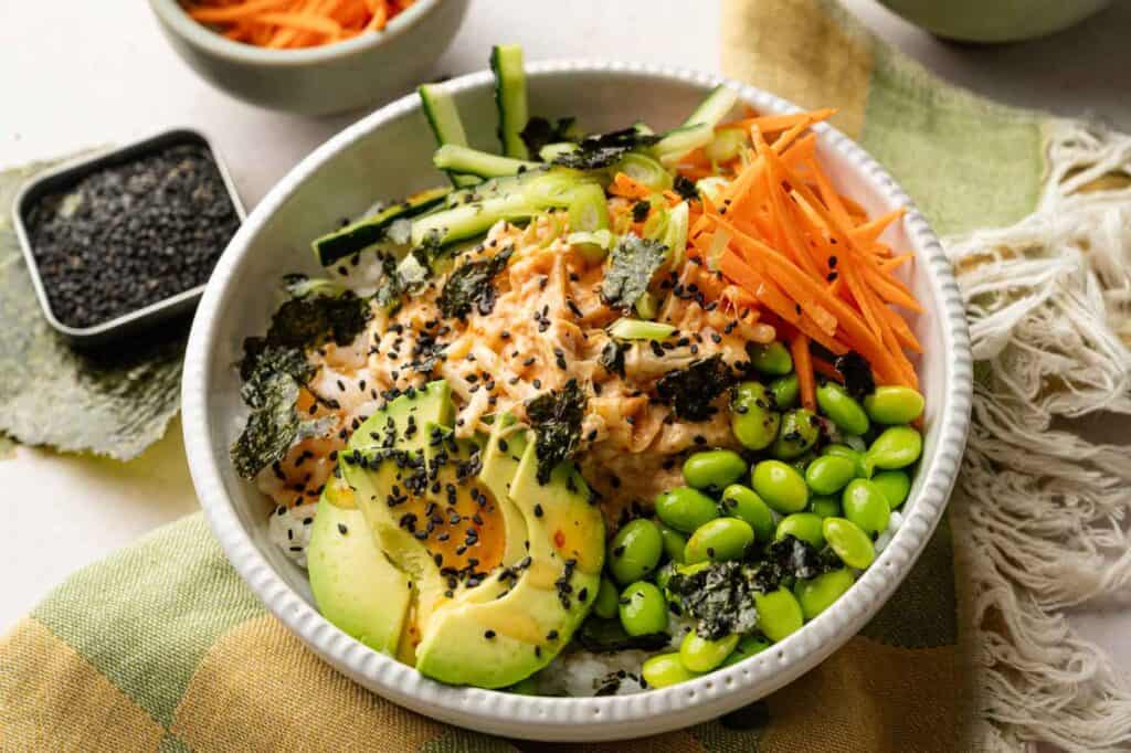 A vibrant Sushi Bowl featuring a bed of rice topped with sliced avocado, shredded carrots, cucumber, edamame, seaweed, black sesame seeds, and a creamy sauce.