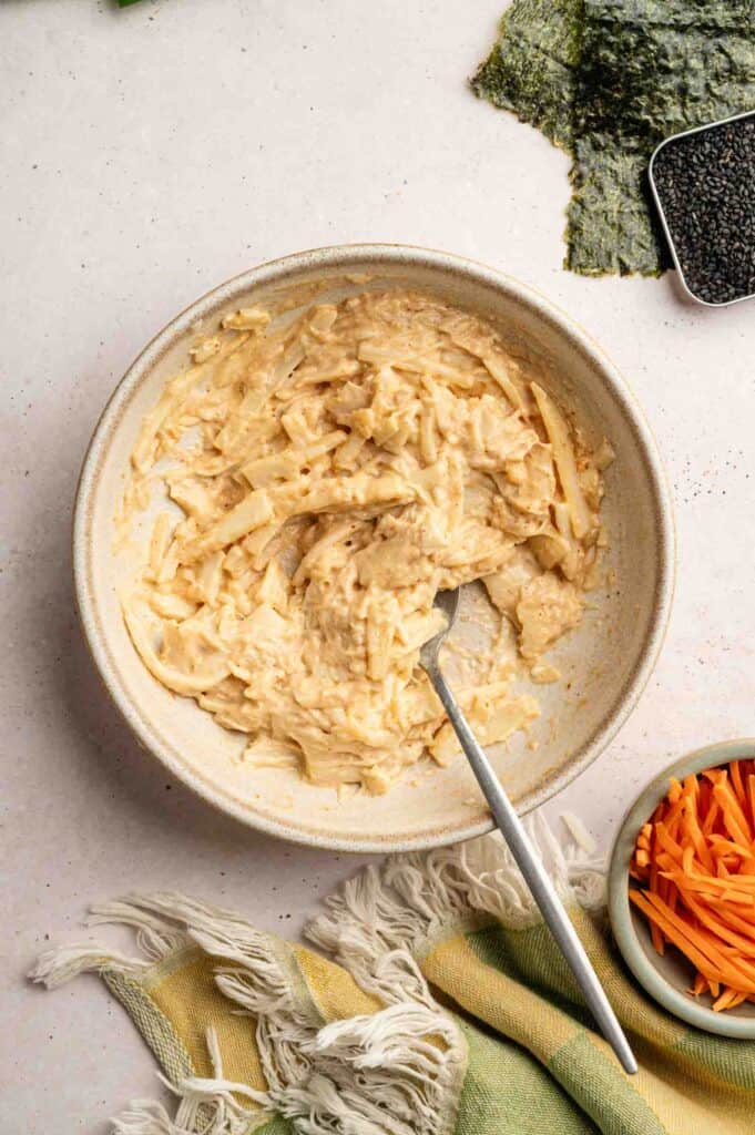 A bowl with spicy tuna mixture sits with a spoon inside, surrounded by sheets of seaweed, black sesame seeds, shredded carrots, and a cloth napkin.