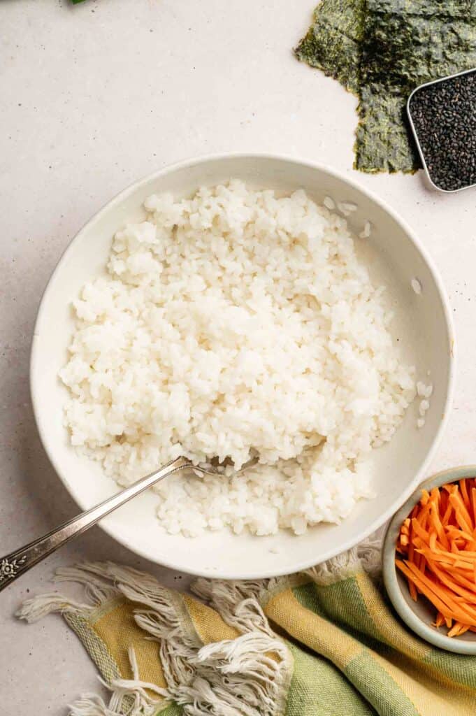 A bowl featuring cooked white rice with a spoon, surrounded by seaweed sheets, black sesame seeds, and a small bowl of shredded carrots.
