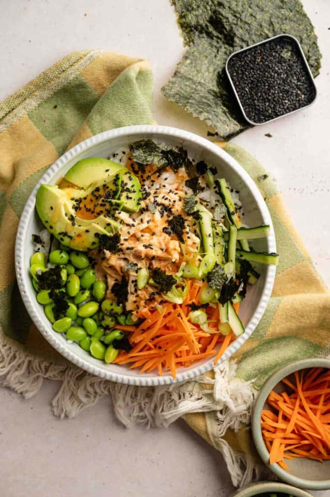 A vibrant Sushi Bowl featuring sliced avocado, shredded carrots, edamame, cucumber, shredded chicken, green onions, seaweed pieces, and black sesame seeds rests on a green and yellow cloth.