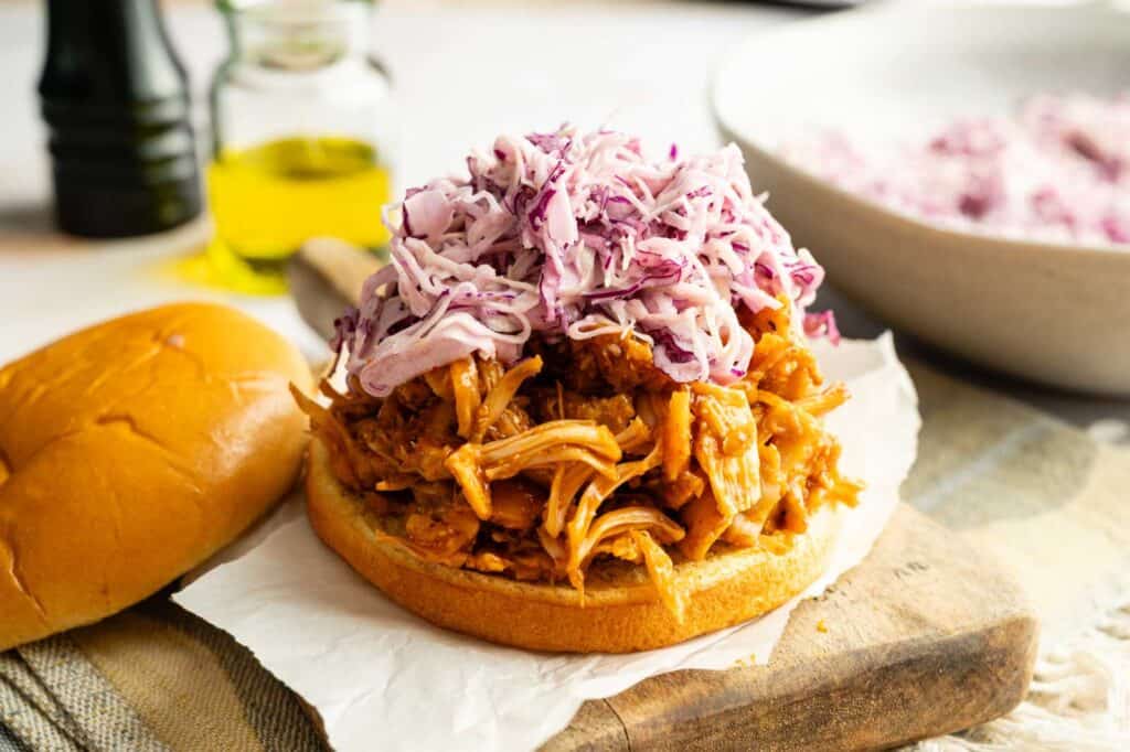 Vegan BBQ Pulled Jackfruit Sandwiches topped with purple cabbage slaw on a bun, next to the top bun, served on a wooden board with a bottle of oil in the background.