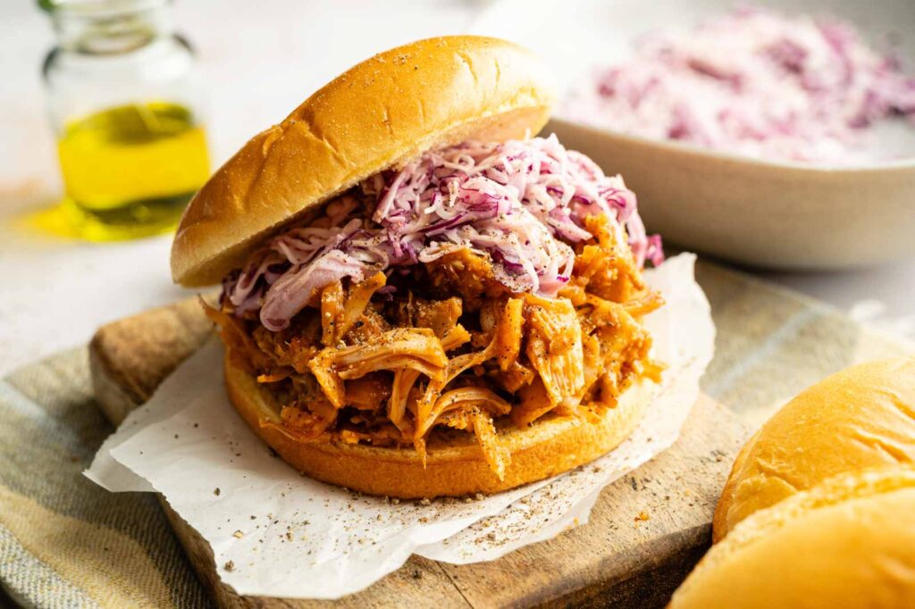 A Vegan BBQ Pulled Jackfruit Sandwich topped with vibrant purple coleslaw sits on a wooden board, accompanied by a bowl of coleslaw and a bottle of oil in the background.