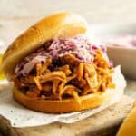 A Vegan BBQ Pulled Jackfruit Sandwich topped with purple cabbage slaw sits on a wooden board, with parchment paper underneath.