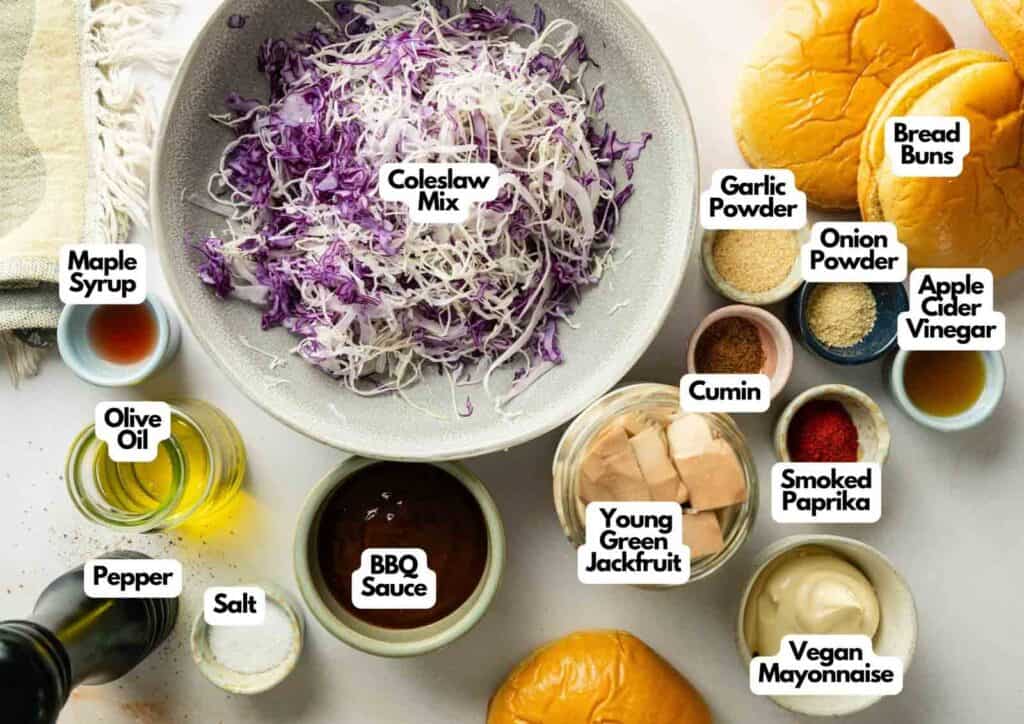 Top-down view of labeled ingredients for Vegan BBQ Pulled Jackfruit Sandwiches, including coleslaw mix, jackfruit, buns, spices, olive oil, BBQ sauce, vegan mayo, and condiments.