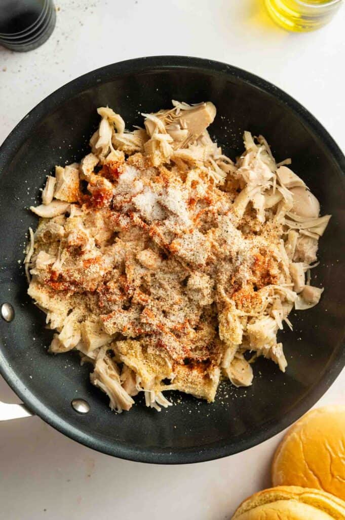Shredded jackfruit in a black skillet, topped with various spices and seasonings, is ready to be piled onto a sandwich bun for delicious Vegan BBQ Pulled Jackfruit Sandwiches.