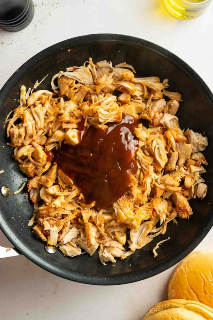 Shredded cooked jackfruit in a skillet topped with barbecue sauce, with a sandwich bun visible in the corner—perfect for those who love classic flavors or crave Vegan BBQ Pulled Jackfruit Sandwiches.