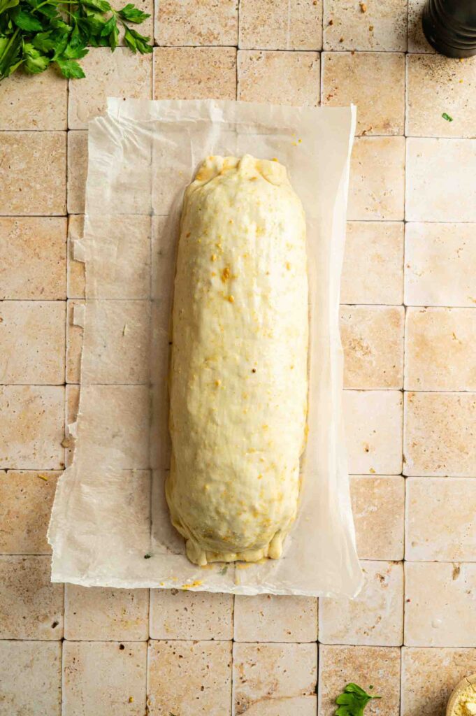 A raw, unbaked Vegan Wellington pastry roll sits on parchment paper atop a tiled surface, with scattered herbs around the edges.