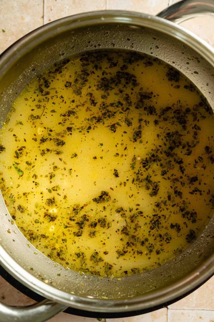 A stainless steel pot filled with hot gravy, with herbs and seasonings floating on the surface, sits on a tiled countertop, ready to accompany a freshly baked Vegan Wellington.