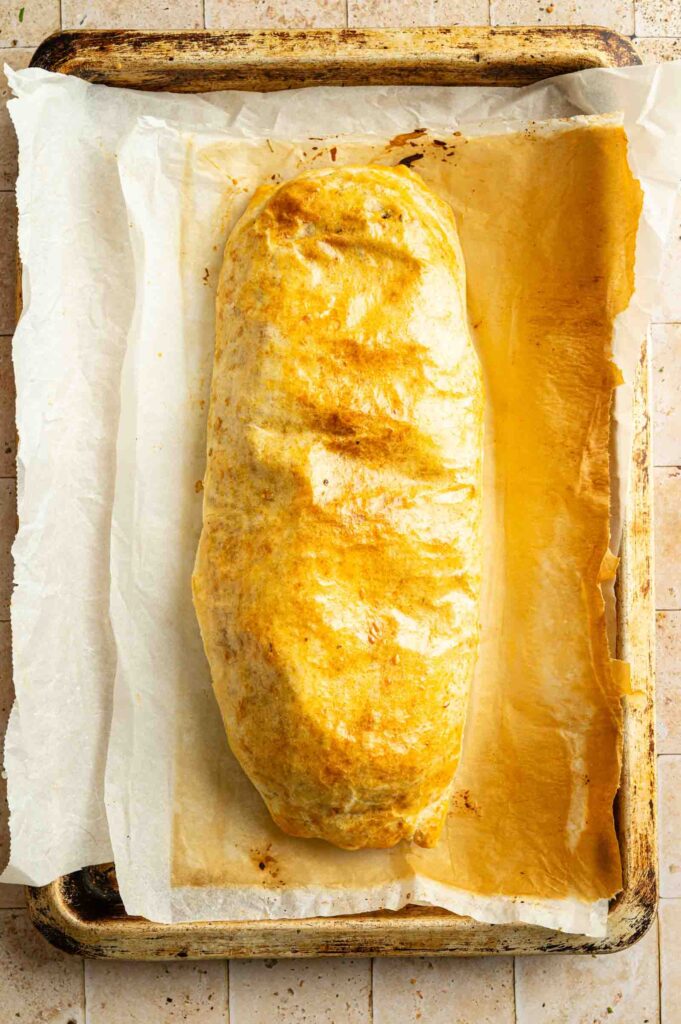 A golden-brown baked Vegan Wellington loaf rests on parchment paper atop a metal baking sheet.
