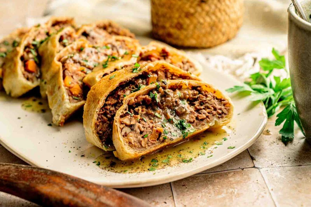 Sliced Vegan Wellington pastry roll filled with a hearty mixture of lentils and vegetables, garnished with chopped herbs on a beige plate.