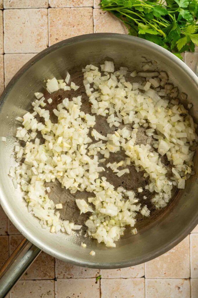 Chopped onions being saut&eacute;ed in a stainless steel pan on a tiled surface, with fresh herbs in the corner&mdash;perfect for starting a flavorful Vegan Wellington.