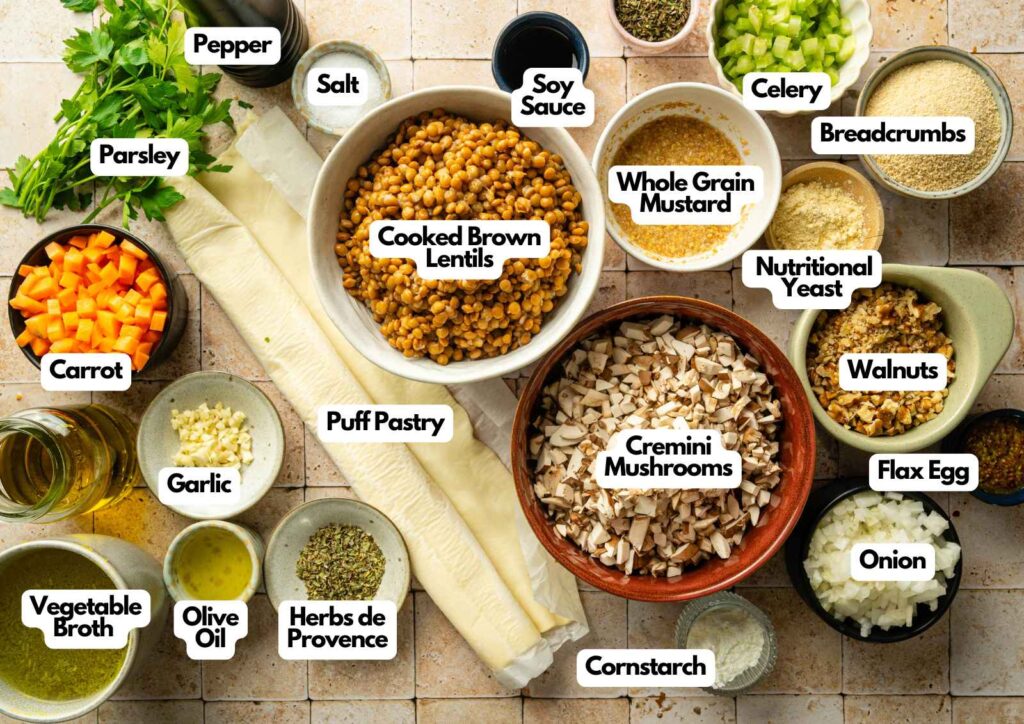 Various labeled ingredients for a Vegan Wellington, including lentils, mushrooms, puff pastry, vegetables, seasonings, oil, broth, and other plant-based items arranged on a countertop.