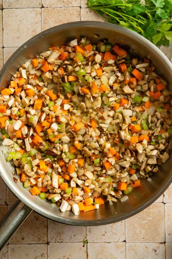 Diced carrots, celery, onions, and mushrooms cooking in a stainless steel pan on a tiled countertop&mdash;perfect savory filling for a Vegan Wellington, with fresh parsley nearby.