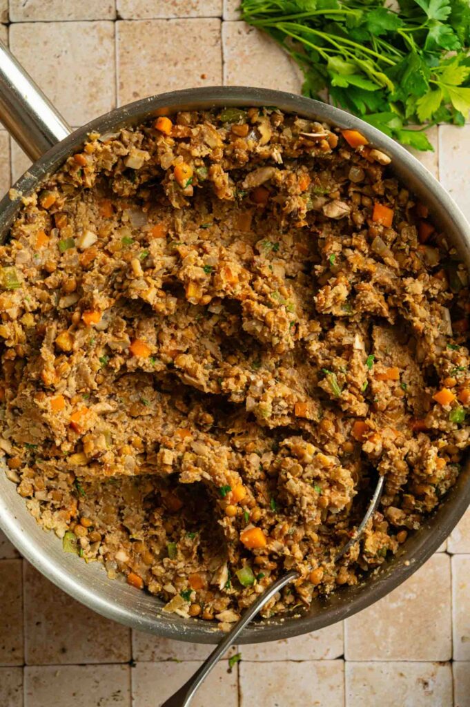 A skillet filled with a cooked mixture of diced vegetables and lentils, reminiscent of a Vegan Wellington filling, with a spoon resting inside. Fresh parsley is visible in the top right corner.